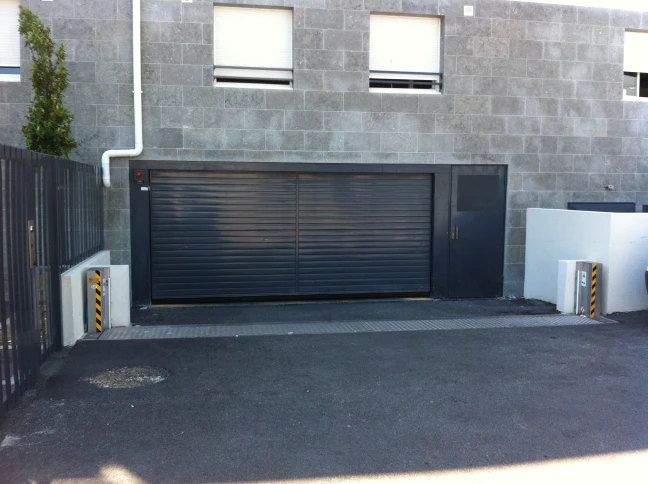 The image shows an entrance to a parking area. There is a closed, black roller door entrance, also with black and yellow striped safety beacons, surrounded by a gray brick wall. The driveway is designed for vehicles and has safety precautions in place.